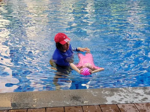 Coach Jina in a red cap supporting a girl in a back float while she holds a ball