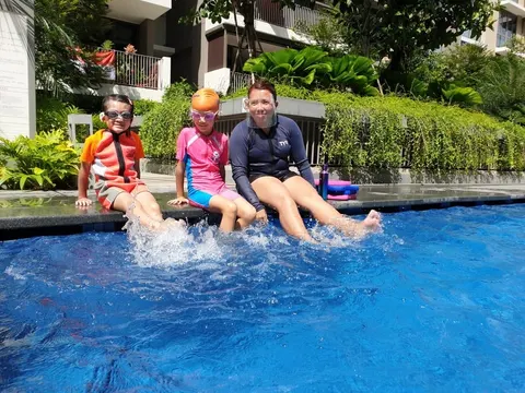 Coach Jina sitting at the pool edge with two smiling kids splashing their legs