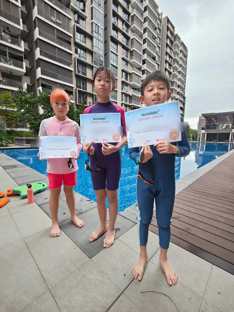 Three children at a condo pool holding their SwimSafer Bronze Award certificates (names blurred)