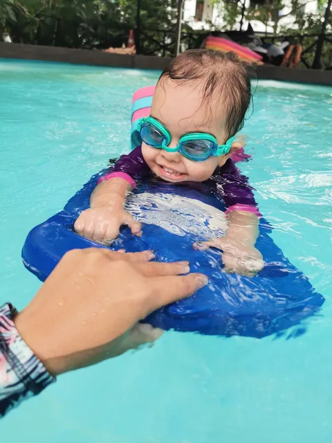 A smiling toddler in goggles on a blue kickboard, steadied by Coach Jina’s hand